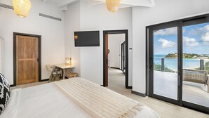 Een slaapkamer met een groot bed, een bureau, een stoel en een televisie. Er is een glazen schuifdeur naar een balkon met uitzicht op zee.
