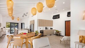 Een moderne keuken met houten kastjes, een eethoek met rieten stoelen, en een groot raam met uitzicht op groen.