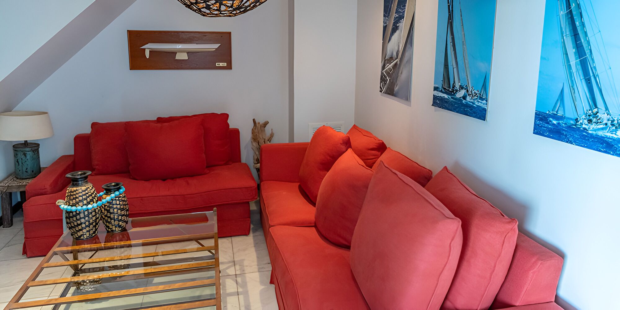 A living room with a red sofa, a glass coffee table, and a lamp. There are wall art pieces and a ceiling light fixture.