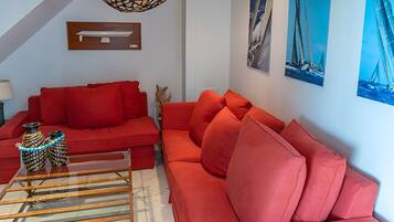 A living room with a red sofa, a glass coffee table, and a lamp. There are wall art pieces and a ceiling light fixture.