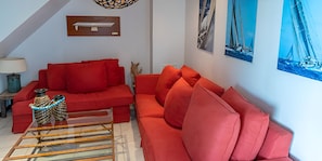 Ein Wohnzimmer mit einem roten Sofa, einem Glastisch und einer Lampe. An den Wänden befinden sich Kunstwerke und eine Deckenleuchte.