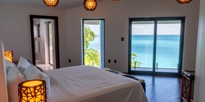 A bedroom with a large bed, two doors leading to a balcony with a view of the sea, and a hanging light fixture. - Your Ideal Retreat Awaits! Coastal Retreat with Ocean Views and Private Pool! (Urlings)
