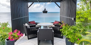A covered outdoor seating area with a view of the sea, featuring wicker furniture and potted plants. - Your Ideal Retreat Awaits! Coastal Retreat with Ocean Views and Private Pool! (Urlings)