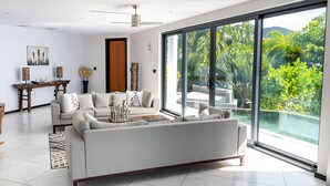 Una sala de estar moderna con un sofá grande, una mesa de centro y una zona de comedor con una mesa y sillas de madera. Hay un ventanal con vistas a la vegetación.