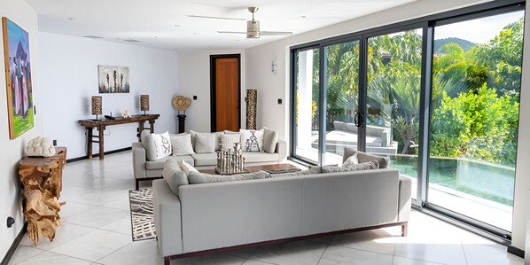 Una sala de estar moderna con un sofá grande, una mesa de centro y un comedor con mesa y sillas de madera. Hay un ventanal con vistas a zonas verdes.