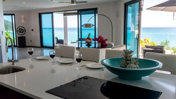 Una cocina moderna con una isla grande, un comedor con vistas al océano, y una mesa de comedor dispuesta con copas de vino y platos.
