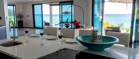 Una cocina moderna con una isla grande, un comedor con vistas al océano, y una mesa de comedor dispuesta con copas de vino y platos.