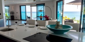 A modern kitchen with a large island, a dining area with a view of the ocean, and a dining table set with wine glasses and plates. - Ocean Blue: Luxury 3-Bedroom Oceanfront Villa with Dual Beach Boardwalk, Antigua (Urlings)