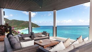 A covered outdoor seating area with wicker furniture and a wooden coffee table, overlooking a beach and ocean. - A Memorable Stay Awaits You Here! Villa Overlooking the Caribbean Sea! (Urlings)