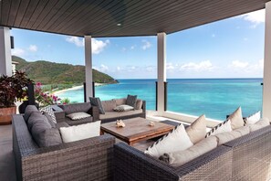 A covered outdoor seating area with wicker furniture and a wooden coffee table, overlooking a beach and ocean. - A Memorable Stay Awaits You Here! Villa Overlooking the Caribbean Sea! (Urlings)