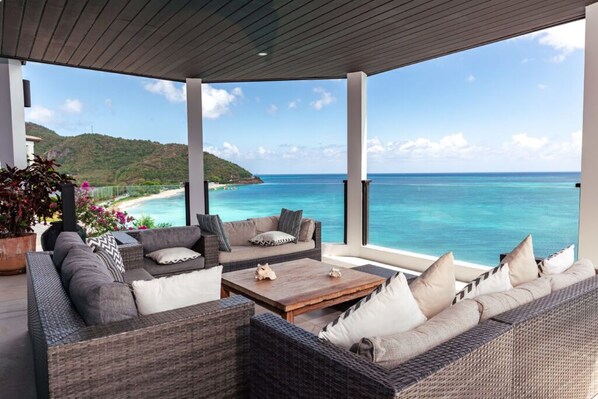 Un espacio de estar al aire libre techado con mobiliario de mimbre y una mesa de centro de madera, con vista a una playa y al océano.