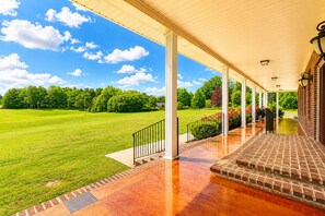 A covered porch with a brick floor, white columns, and a view of a green lawn and trees.