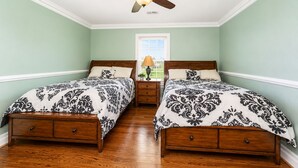 A bedroom with two wooden beds, a ceiling fan, a window with blinds, and a nightstand with a lamp.