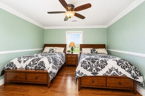 A bedroom with two wooden beds, a ceiling fan, a window with blinds, and a nightstand with a lamp.