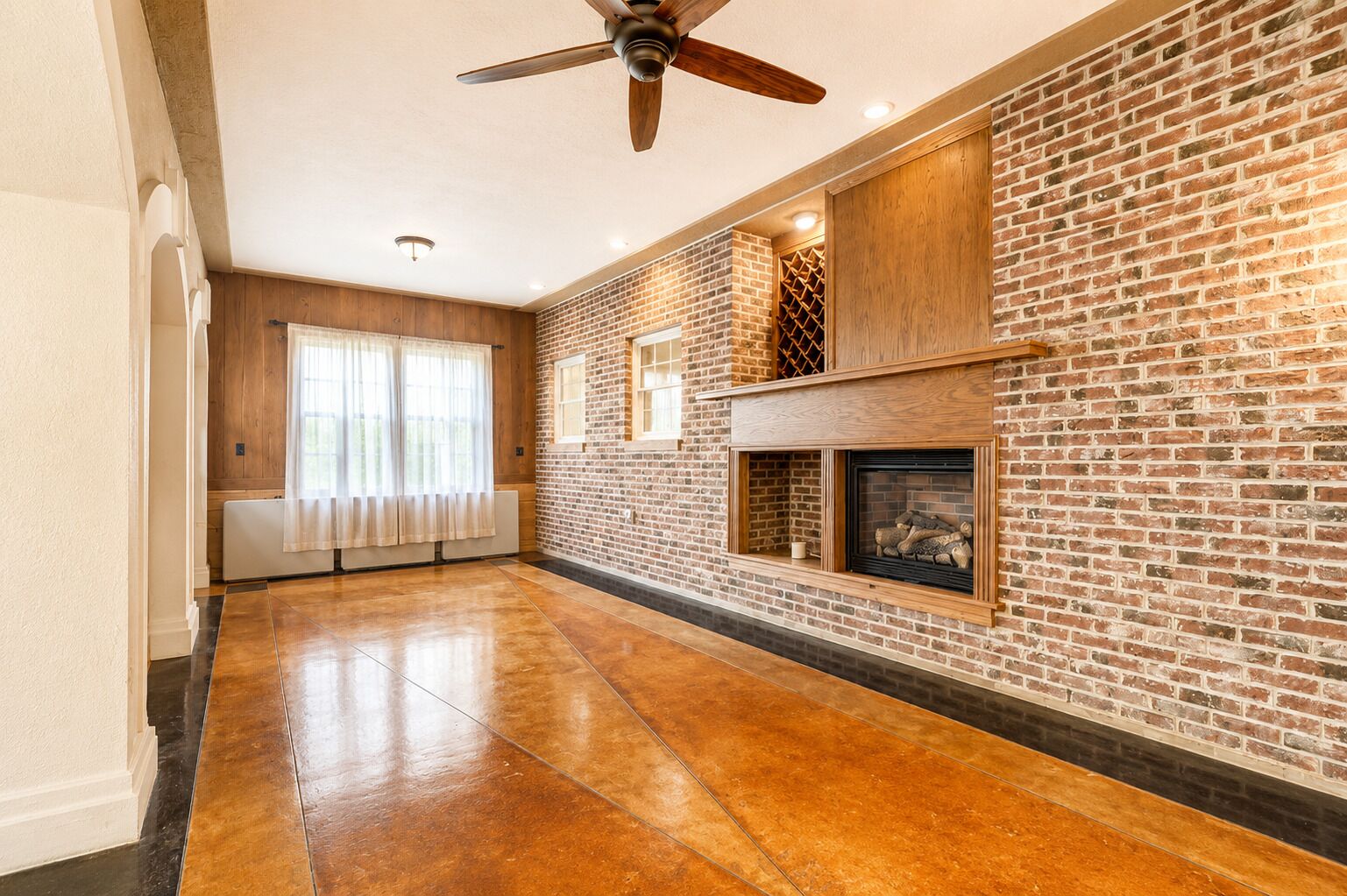 A spacious room with a brick fireplace, a ceiling fan, and a large window with curtains.