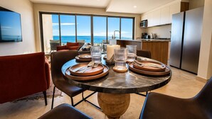 Uma sala de jantar moderna com uma mesa grande posta para quatro, uma cozinha ao fundo e vista para o oceano através de grandes janelas.