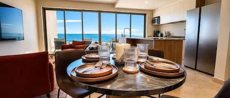 Un comedor moderno con una mesa grande dispuesta para cuatro, una cocina al fondo y vistas al océano a través de amplios ventanales.