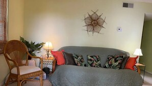 A room with a grey sofa, a wicker chair, two side tables with lamps, a wall decoration, and a plant.