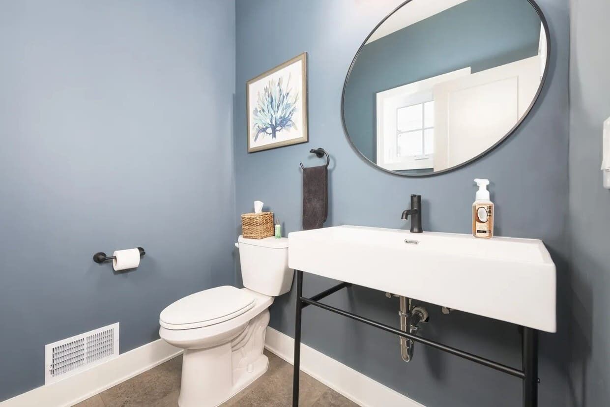 A bathroom with a white toilet, a white sink with a black frame, a round mirror, and a framed artwork on the wall.