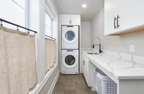 A compact laundry room with a front-loading washer and dryer, a sink, and a laundry basket. - Blue Haven | 5 min walk to beach • pool • grill (South Haven)
