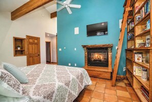 A bedroom with a fireplace, a bed with patterned bedding, a wooden bookshelf, and a television mounted on the wall. - Wildhaus Retreat | Hot Tub • Private Walking Trail (South Haven)