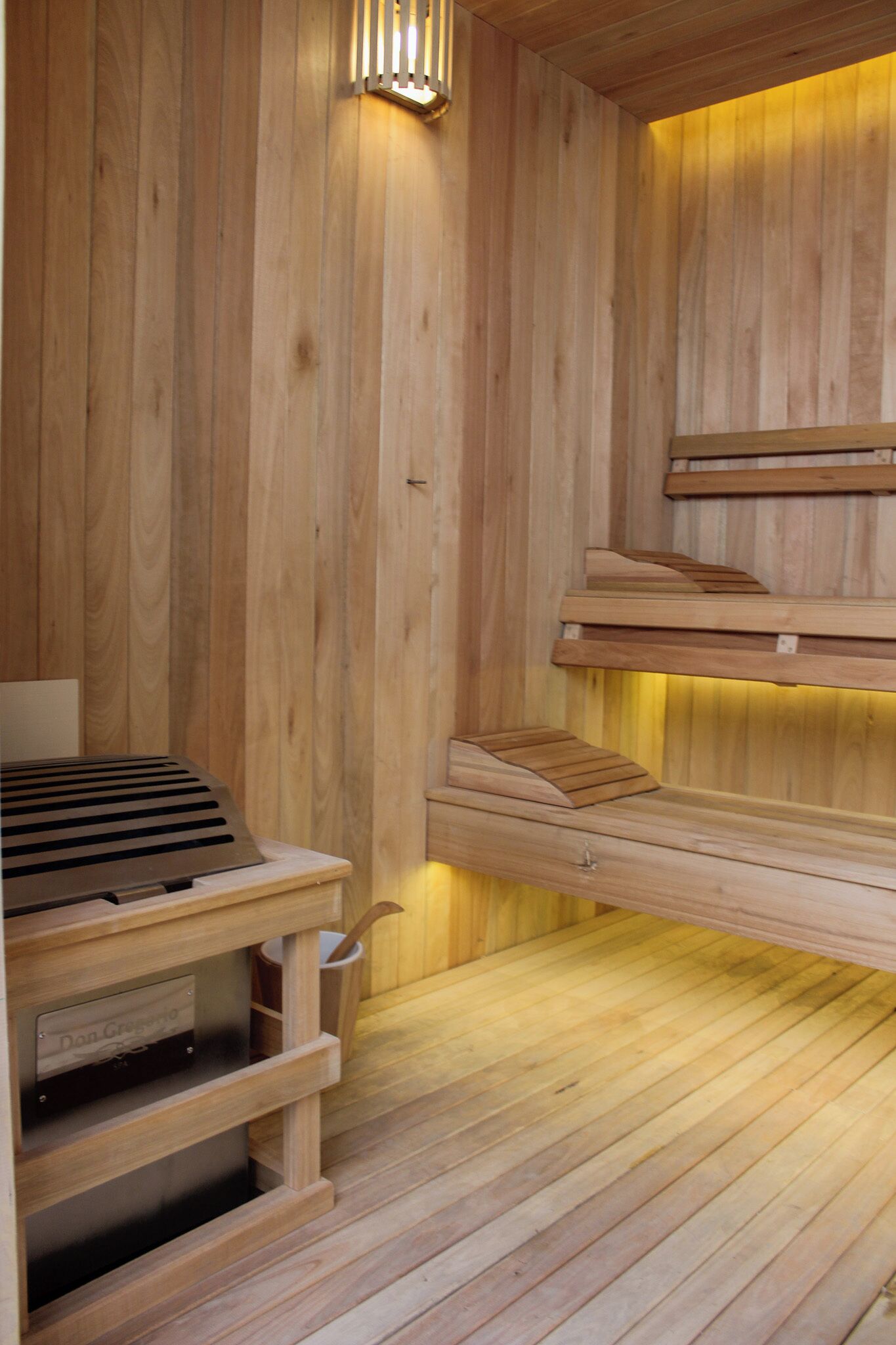 A wooden sauna with benches and a table, featuring a sloped ceiling and a wall-mounted light fixture.