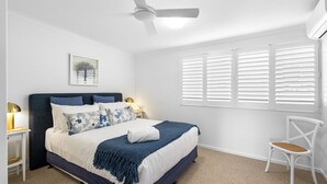 Quarto com cama, mesas de cabeceira, uma cadeira, ventoinha no teto e janela com persianas.