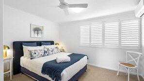 Ein Schlafzimmer mit einem Bett, Nachttischen, einem Sessel, einem Deckenventilator und einem Fenster mit Jalousien.
