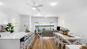 A modern kitchen with white cabinetry, a central island, and a dining area with white chairs. - The Anchorage 7 Noosaville (Noosaville)