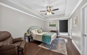 Ein Schlafzimmer mit Bett, einem Sessel, einem Deckenventilator, einem Gemälde und einem Fenster.