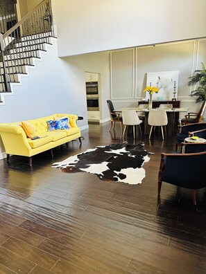 Ein modernes Wohnzimmer mit einem gelben Sofa, einem Esstisch mit Stühlen, einem Kuhfellteppich und einer Treppe mit Metallgeländern.