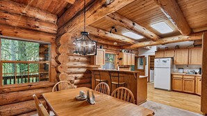 A log cabin interior with a dining area, kitchen, and large windows.