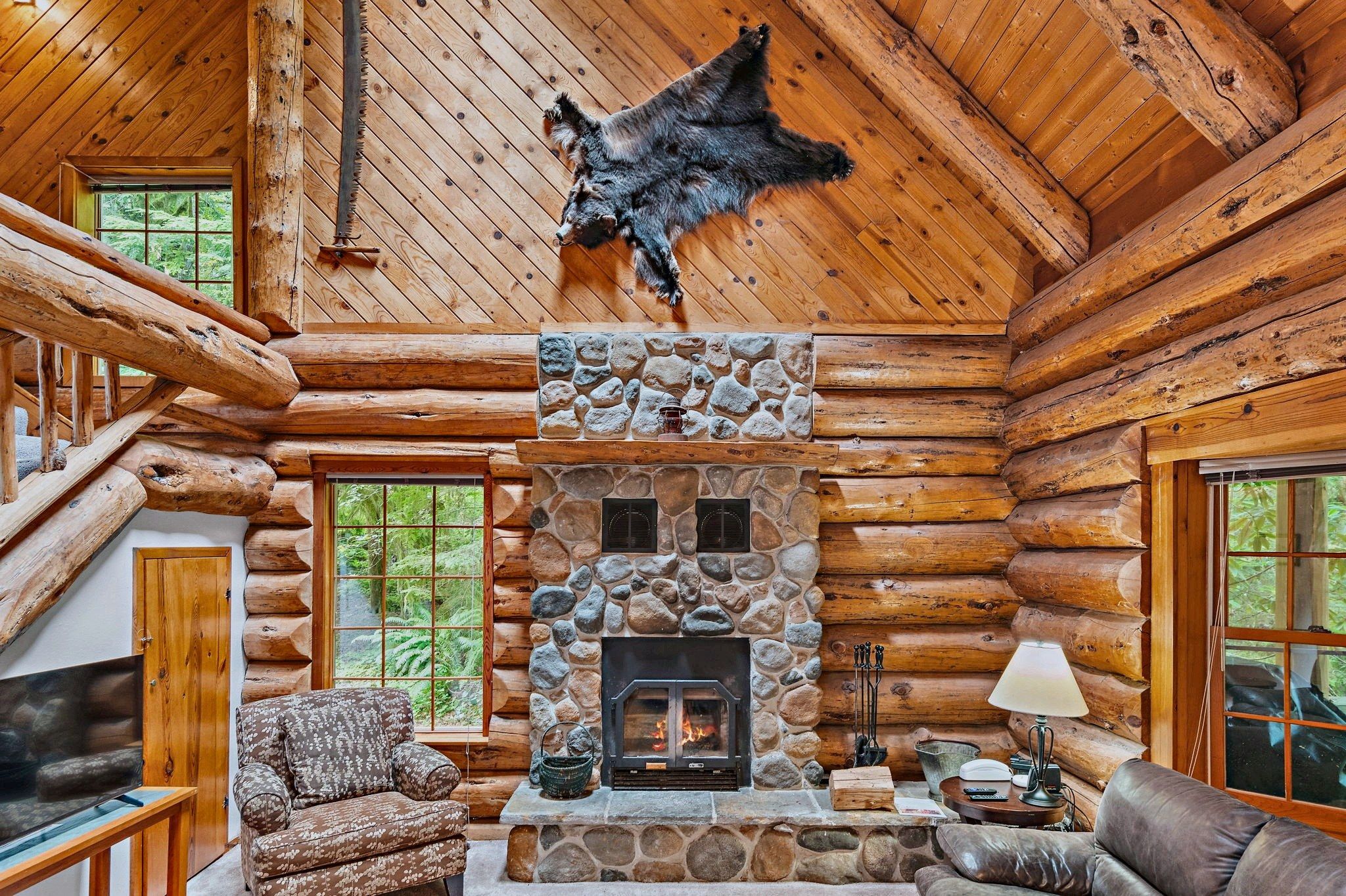 A log cabin interior with a stone fireplace, wooden walls, and a bear hide on the ceiling.