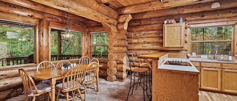 A log cabin interior with a dining area, kitchen, and large windows offering views of the forest.