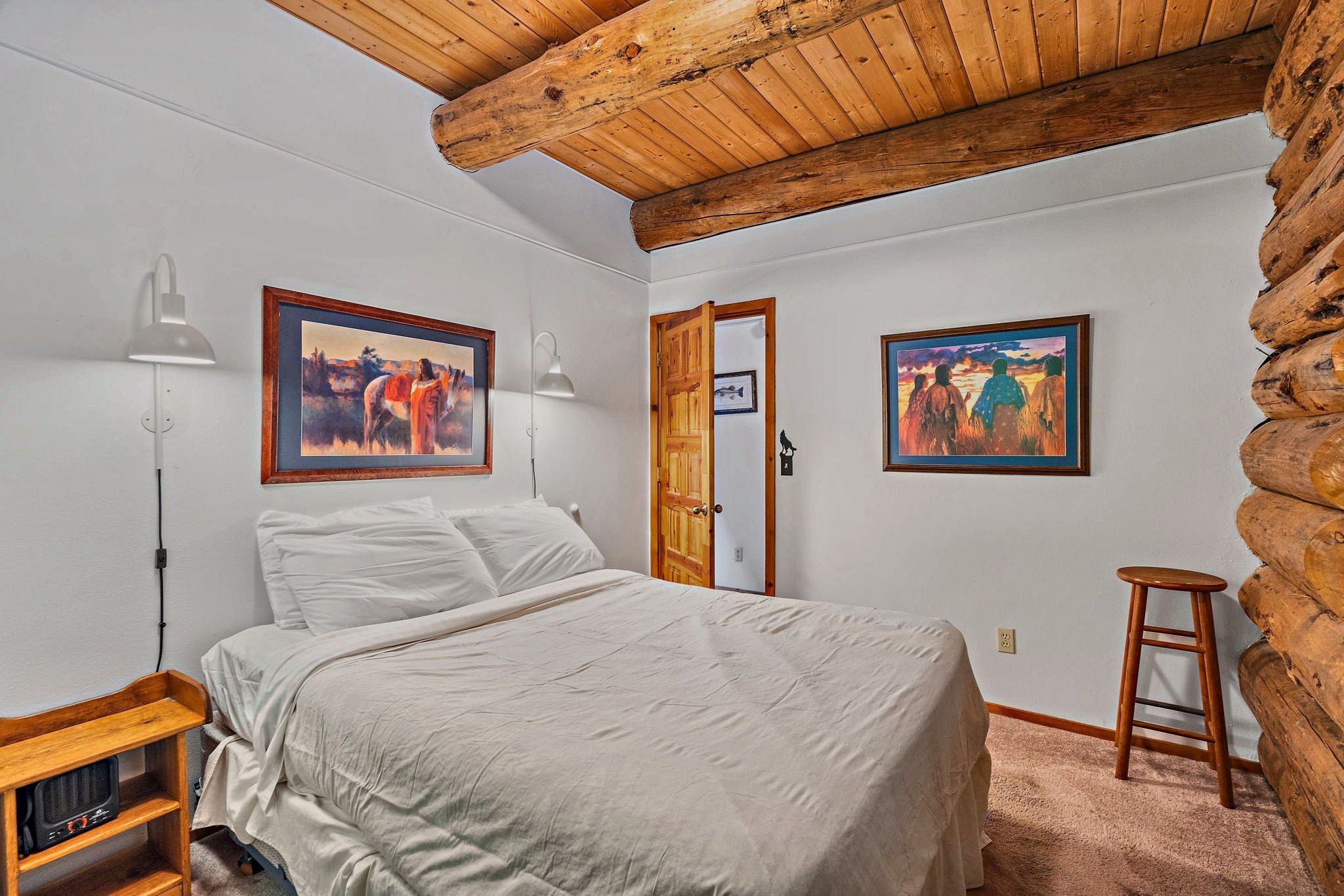 A bedroom with a bed, two wall-mounted lamps, a wooden door, and two framed paintings.