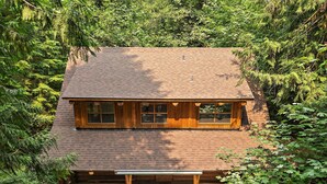A two-story wooden cabin with a porch, surrounded by dense forest.