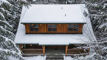 A two-story wooden cabin with a snow-covered roof, surrounded by dense forest.