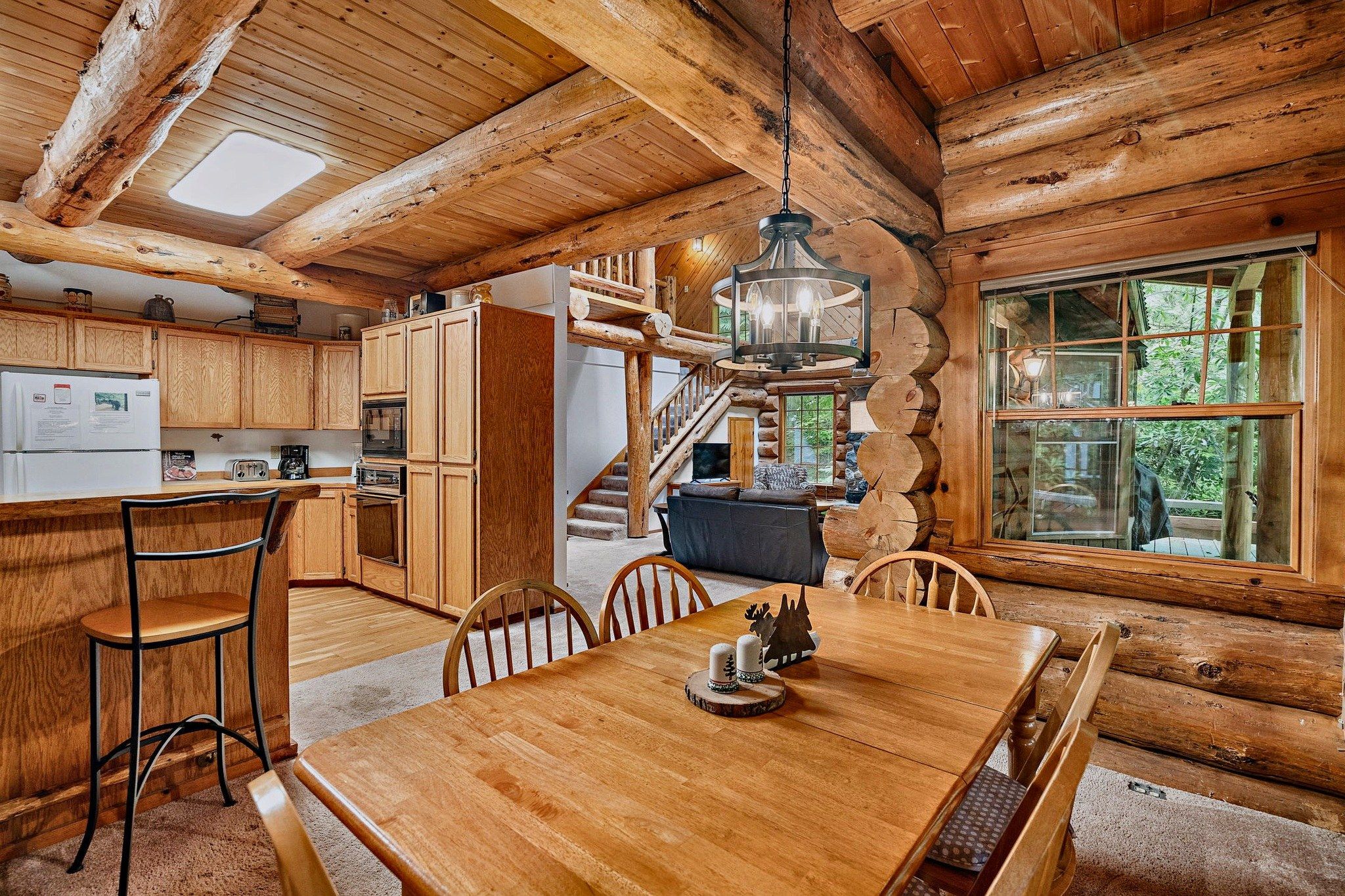 Interior rústico de una cabaña de troncos con una mesa de comedor de madera, sillas, una zona de cocina con electrodomésticos y una escalera que sube a un nivel superior.