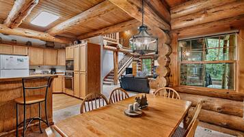 Interior rústico de una cabaña de troncos con una mesa de comedor de madera, sillas, una zona de cocina con electrodomésticos y una escalera que sube a un nivel superior.