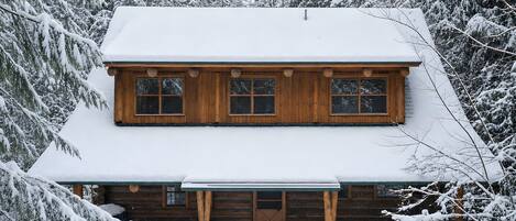 Una cabaña de troncos de dos pisos con techo cubierto de nieve y un porche delantero.