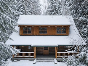 A two-story log cabin with a snow-covered roof and a front porch.