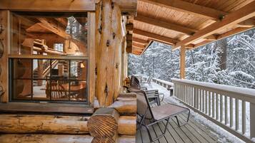 A wooden cabin with a covered porch, a dining area with chairs, and a view of snow-covered trees.