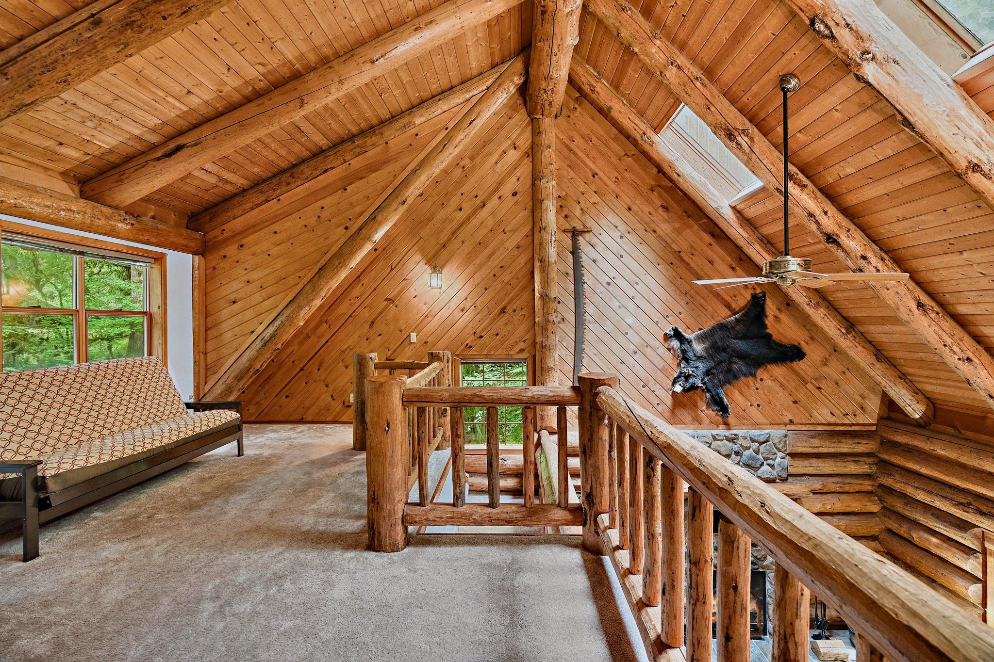 Interior de una cabaña de troncos con un entrepiso que funciona como dormitorio, una cama de madera, una chimenea de piedra y un oso montado en la pared.