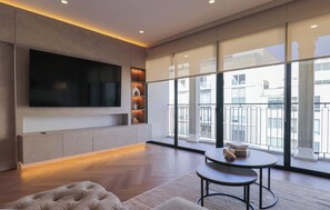 Ein modernes Wohnzimmer mit einem großen Flachbildfernseher, einer Couch, einem Couchtisch und Blick auf die Stadt durch hohe Fenster.