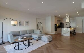 Ein modernes Wohnzimmer mit einem grauen Sofa, einem runden Couchtisch und einem weißen Hocker. Im Hintergrund ist eine Küchenzeile mit einem Esstisch und Stühlen zu sehen.