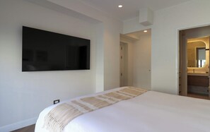 Ein Schlafzimmer mit einem großen Bett, einem flachbildfernseher an der Wand und einem durchgehenden Türspalt einsehbaren Badezimmer.