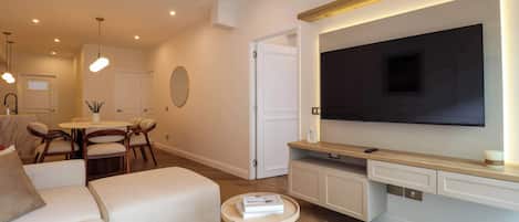 A modern living room with a flat-screen TV, a sofa, a coffee table, and a dining area with chairs and a table.