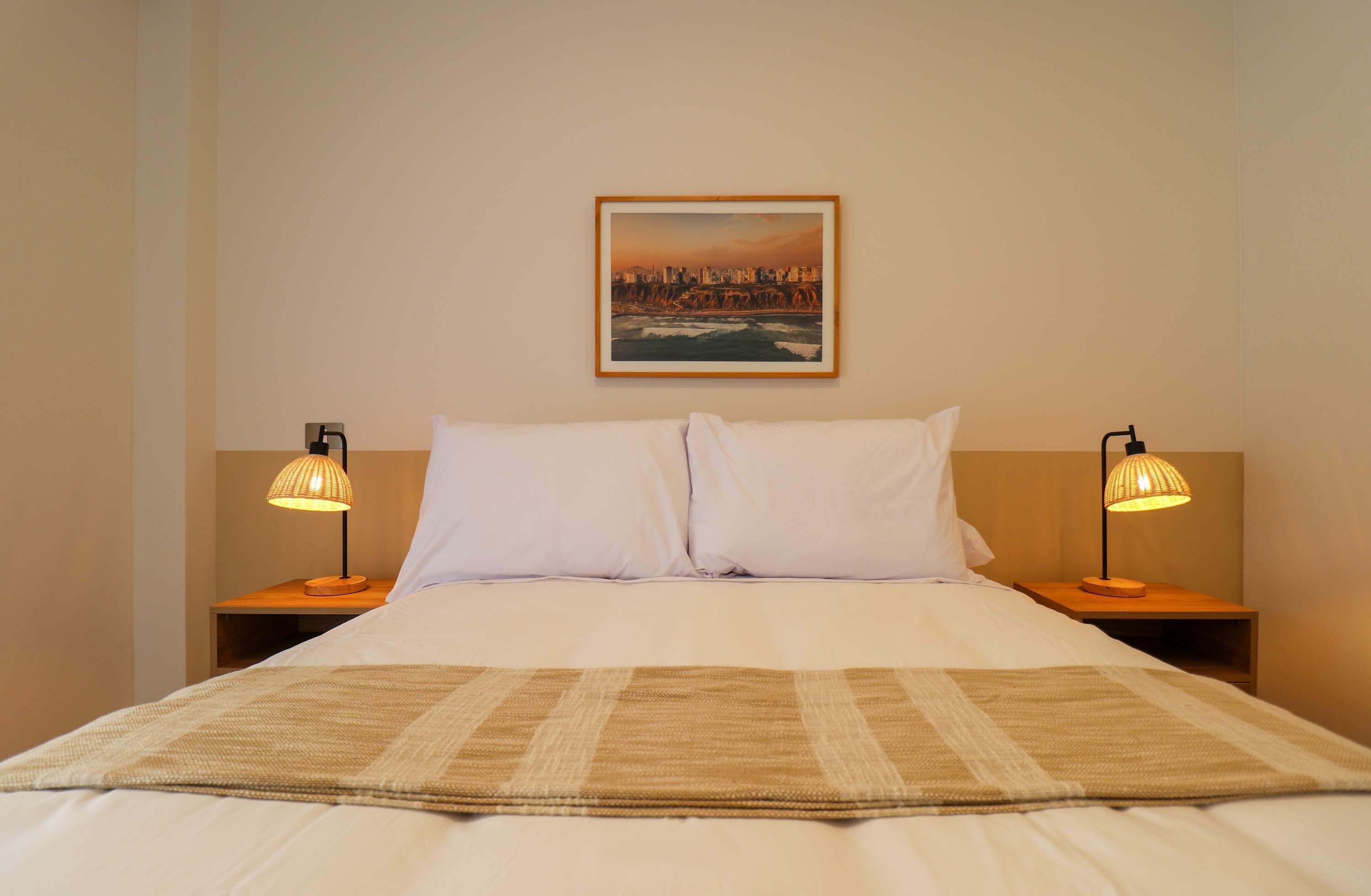 Une chambre d’hôtel avec un lit, deux lampes de chevet, un tableau représentant un paysage urbain encadré au mur et un plaid à motifs.