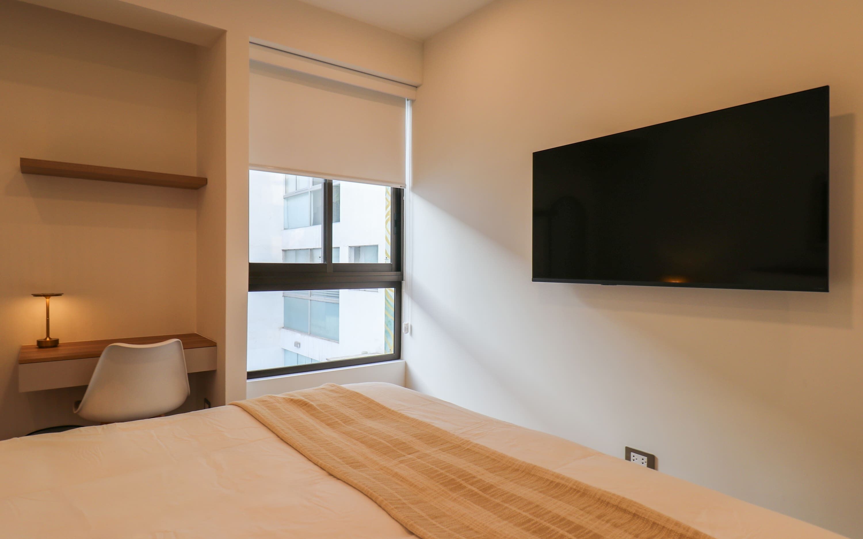 A hotel room with a bed, a bedside table with a lamp, a TV mounted on the wall, and a window with a view of buildings.