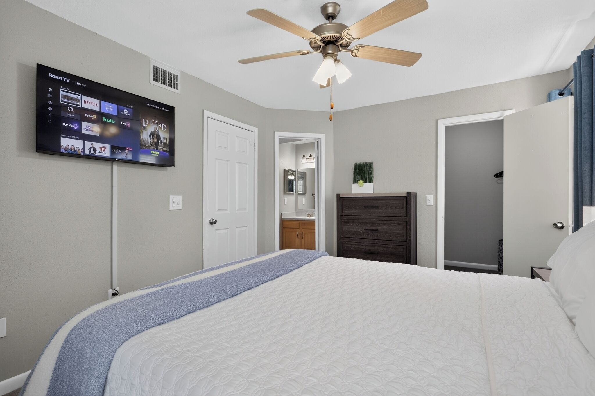 Une chambre à coucher comprenant un lit, un ventilateur de plafond, un téléviseur fixé au mur et une porte donnant accès à une salle de bain.